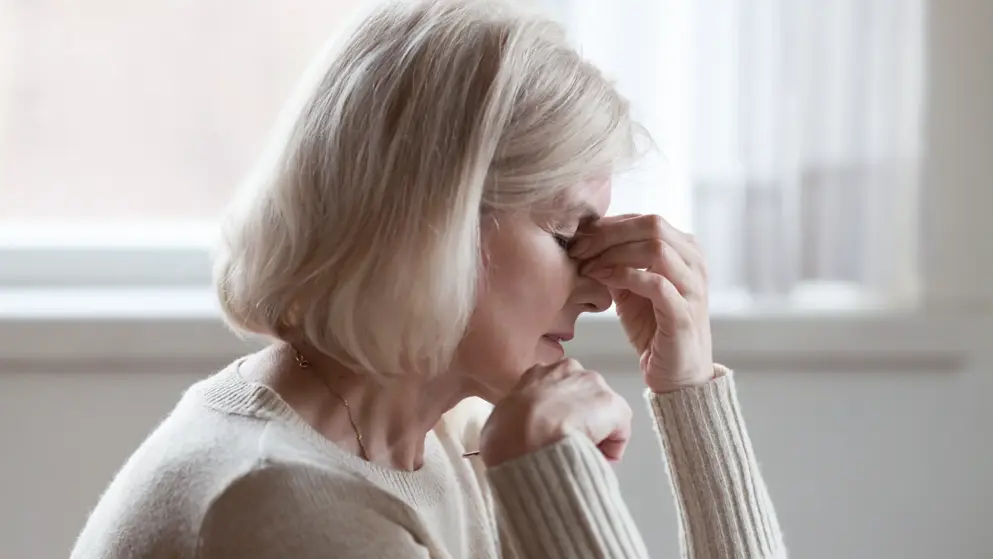 Fatigued upset middle aged older woman massaging nose bridge feeling eye strain or headache trying to relieve pain. Woman has blond hair and cream jumper, white background