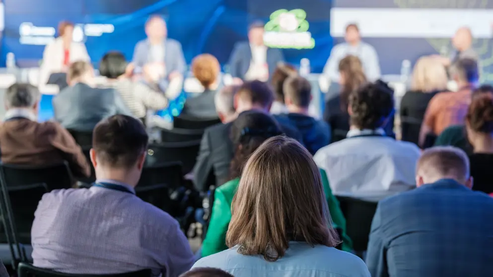 Focused audience members view a panel discussion at a conference. The image captures engagement and diverse professionals in a blurred background.